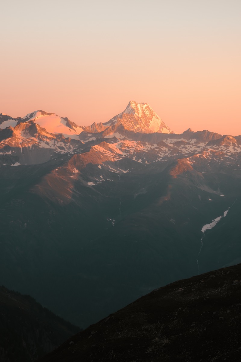 Schneebedeckter Gipfel im rosa Abendlicht über dunklen Bergrücken