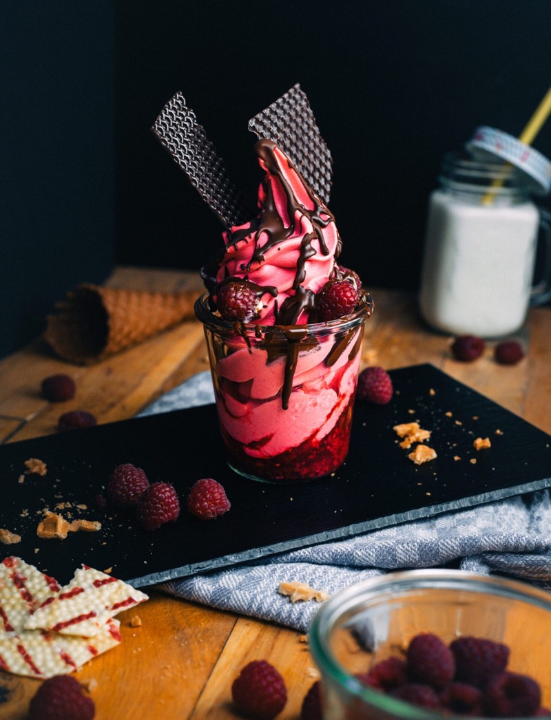 Himbeer-Softeis im Glas mit Schokoladensoße und Waffeln, daneben frische Himbeeren