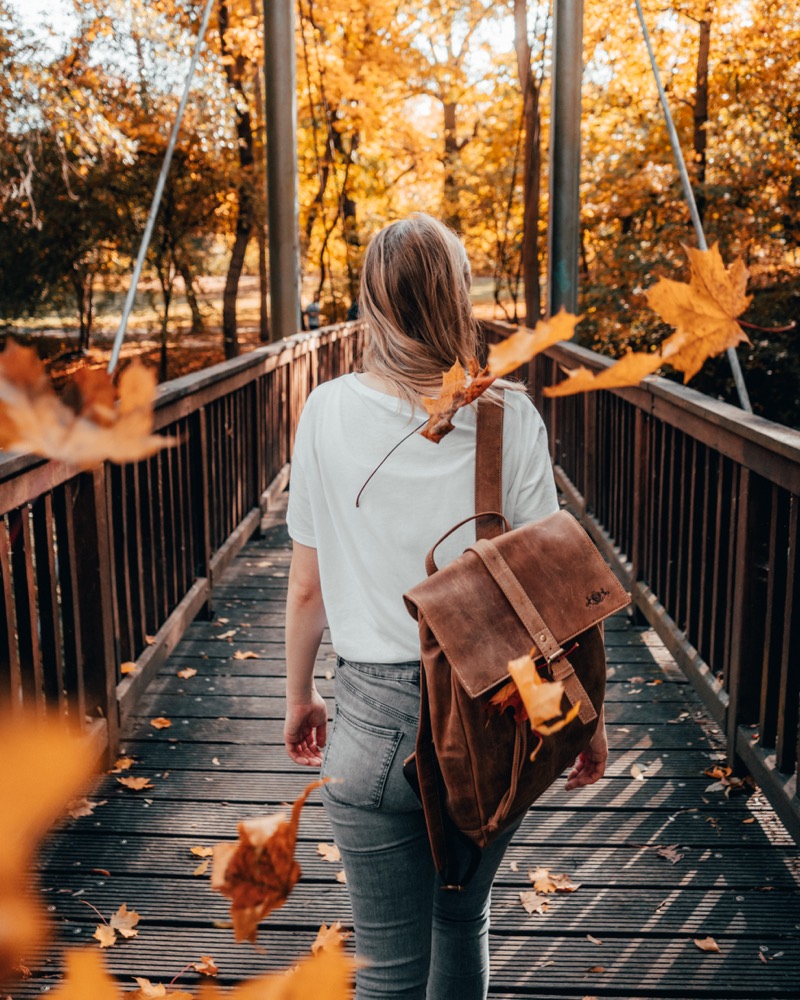 Frau mit Ledertasche auf einem Holzsteg zwischen herbstlich gelben Bäumen