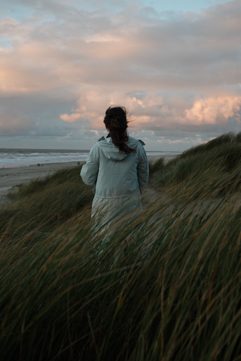 Frau in heller Jacke steht im hohen Gras an einer Steilküste und blickt aufs Meer