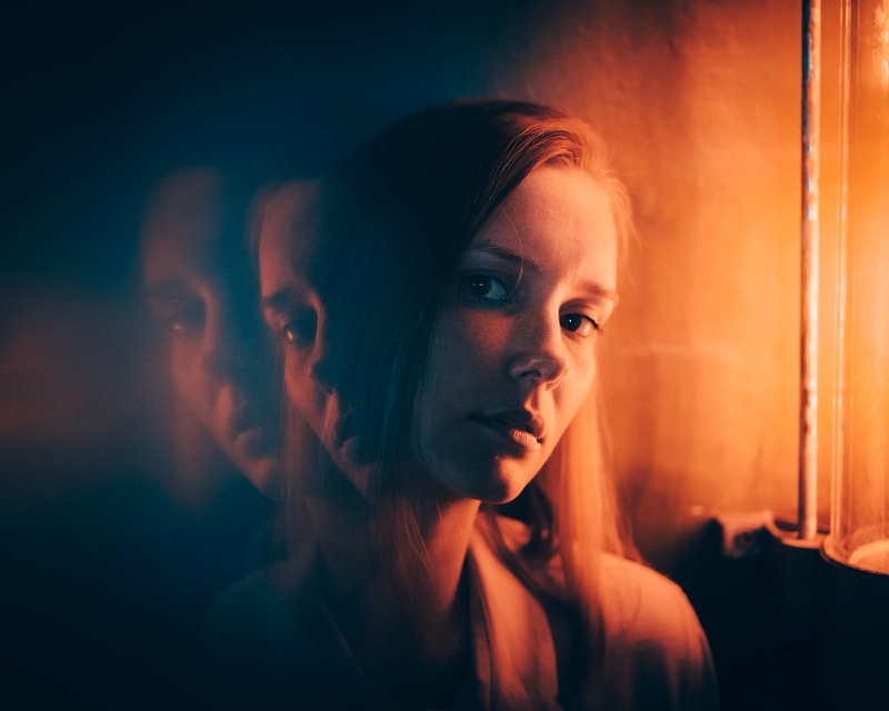 Frau im orangen Licht vor einer Glasscheibe, die ihr Spiegelbild zeigt
