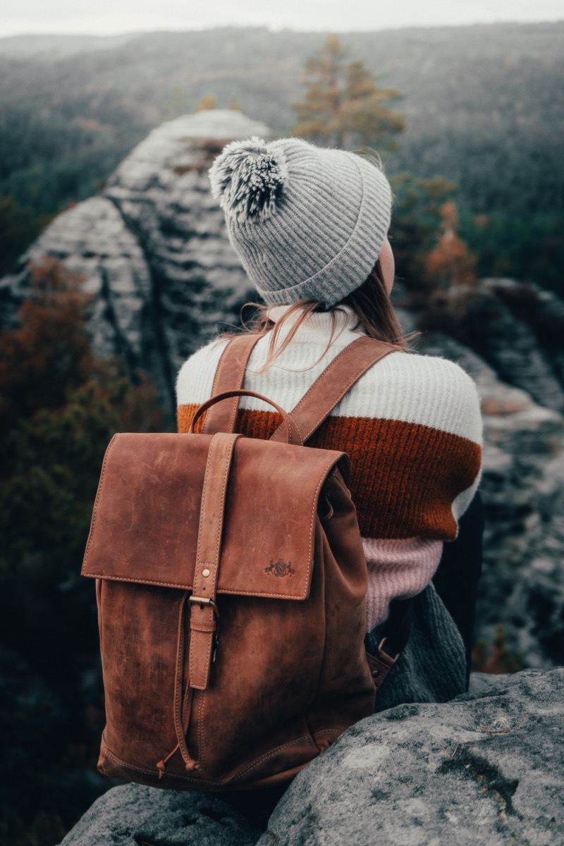 Frau mit Mütze und braunem Lederrucksack sitzt auf einem Felsen über einem Waldtal